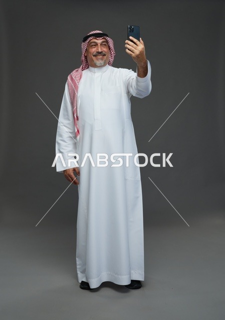 رجل  عربي سعودي خليجي مسن يرتدي الزي الثوب الابيض والشماغ ويمسك الهاتف المحمول, التقاط صورة سيلفي, اجراء مكالمة فيديو عبر الجوال,  ايماءات واشارات مختلفه , كامل الجسم, خلفيه رماديه