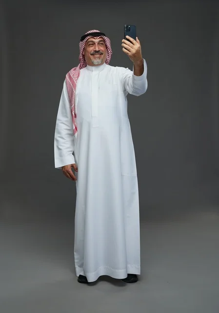 An elderly Arab Saudi Gulf man wearing a white thobe and a shemagh, holding a mobile phone, taking a selfie, making a video call via the phone, with various gestures and signals, full body, gray background.