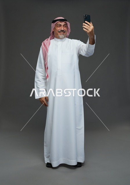 رجل  عربي سعودي خليجي مسن يرتدي الزي الثوب الابيض والشماغ ويمسك الهاتف المحمول, التقاط صورة سيلفي, اجراء مكالمة فيديو عبر الجوال,  ايماءات واشارات مختلفه , كامل الجسم, خلفيه رماديه