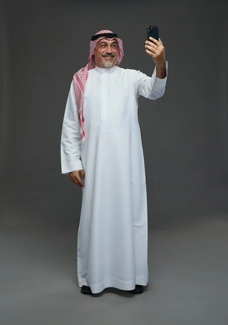 رجل  عربي سعودي خليجي مسن يرتدي الزي الثوب الابيض والشماغ ويمسك الهاتف المحمول, التقاط صورة سيلفي, اجراء مكالمة فيديو عبر الجوال,  ايماءات واشارات مختلفه , كامل الجسم, خلفيه رماديه