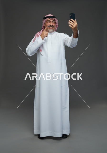 رجل  عربي سعودي خليجي مسن يرتدي الزي الثوب الابيض والشماغ ويمسك الهاتف المحمول, التقاط صورة سيلفي, اجراء مكالمة فيديو عبر الجوال,  ايماءات واشارات مختلفه , كامل الجسم, خلفيه رماديه