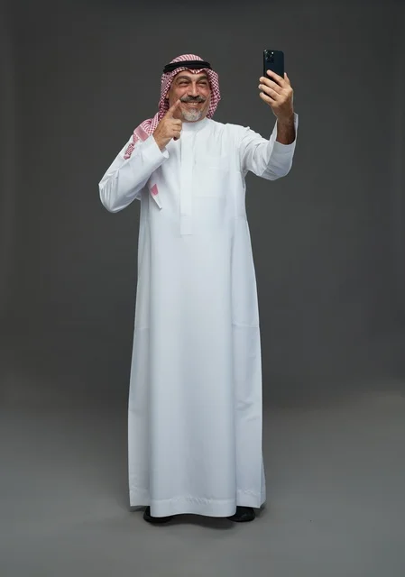 رجل  عربي سعودي خليجي مسن يرتدي الزي الثوب الابيض والشماغ ويمسك الهاتف المحمول, التقاط صورة سيلفي, اجراء مكالمة فيديو عبر الجوال,  ايماءات واشارات مختلفه , كامل الجسم, خلفيه رماديه