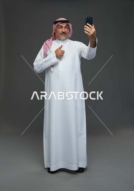 رجل  عربي سعودي خليجي مسن يرتدي الزي الثوب الابيض والشماغ ويمسك الهاتف المحمول, التقاط صورة سيلفي, اجراء مكالمة فيديو عبر الجوال,  ايماءات واشارات مختلفه , كامل الجسم, خلفيه رماديه