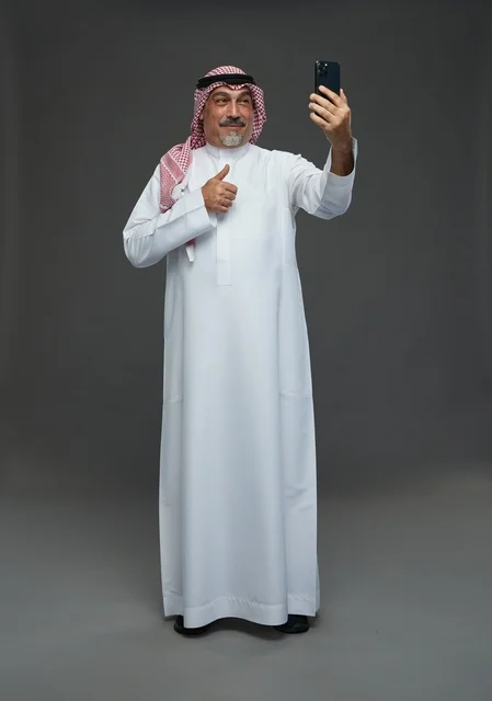 An elderly Arab Saudi Gulf man wearing a white thobe and a shemagh, holding a mobile phone, taking a selfie, making a video call via the phone, with various gestures and signals, full body, gray background.