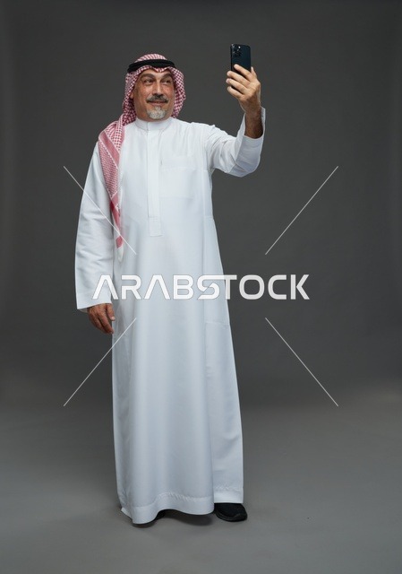 رجل  عربي سعودي خليجي مسن يرتدي الزي الثوب الابيض والشماغ ويمسك الهاتف المحمول, التقاط صورة سيلفي, اجراء مكالمة فيديو عبر الجوال,  ايماءات واشارات مختلفه , كامل الجسم, خلفيه رماديه