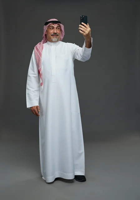 An elderly Arab Saudi Gulf man wearing a white thobe and a shemagh, holding a mobile phone, taking a selfie, making a video call via the phone, with various gestures and signals, full body, gray background.