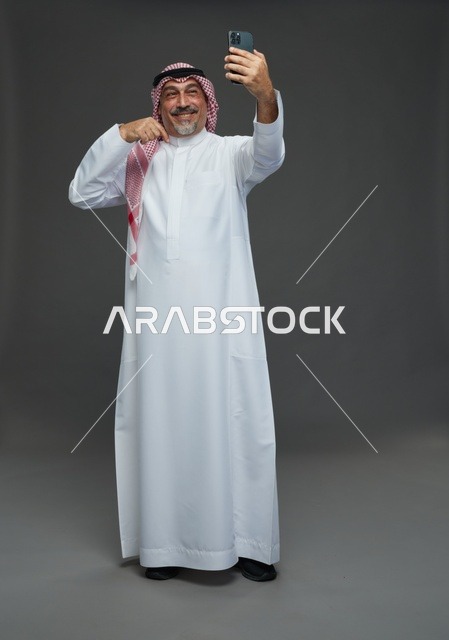 An elderly Arab Saudi Gulf man wearing a white thobe and a shemagh, holding a mobile phone, taking a selfie, making a video call via the phone, with various gestures and signals, full body, gray background.