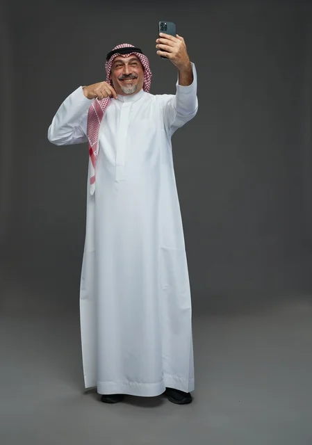 رجل  عربي سعودي خليجي مسن يرتدي الزي الثوب الابيض والشماغ ويمسك الهاتف المحمول, التقاط صورة سيلفي, اجراء مكالمة فيديو عبر الجوال,  ايماءات واشارات مختلفه , كامل الجسم, خلفيه رماديه