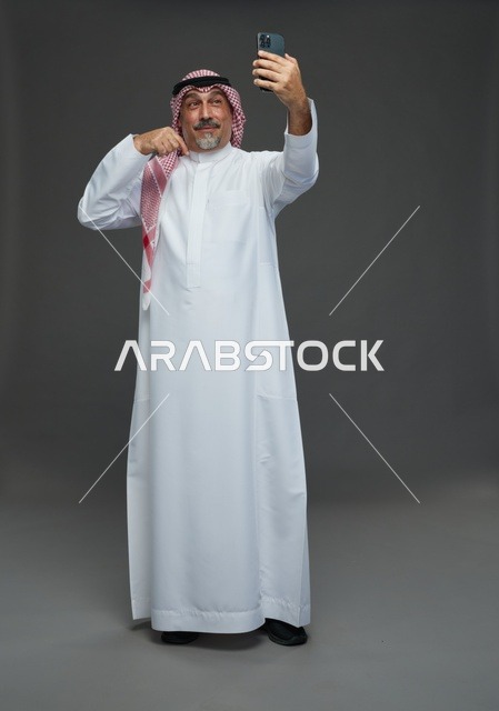 رجل  عربي سعودي خليجي مسن يرتدي الزي الثوب الابيض والشماغ ويمسك الهاتف المحمول, التقاط صورة سيلفي, اجراء مكالمة فيديو عبر الجوال,  ايماءات واشارات مختلفه , كامل الجسم, خلفيه رماديه