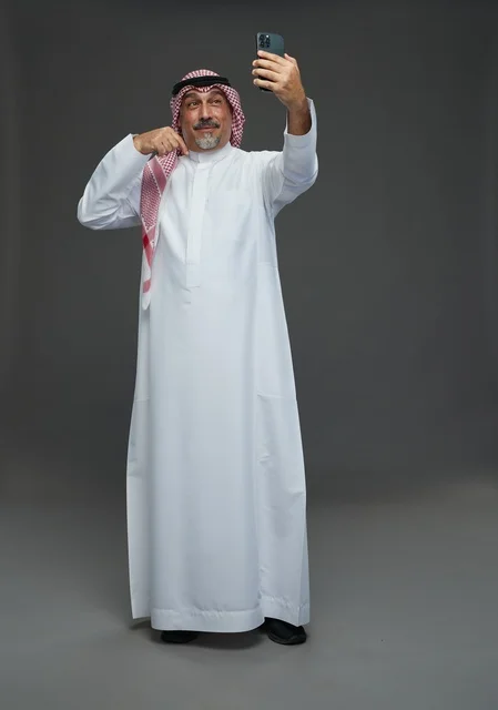 An elderly Arab Saudi Gulf man wearing a white thobe and a shemagh, holding a mobile phone, taking a selfie, making a video call via the phone, with various gestures and signals, full body, gray background.