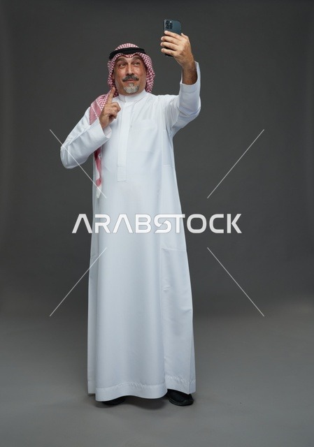 رجل  عربي سعودي خليجي مسن يرتدي الزي الثوب الابيض والشماغ ويمسك الهاتف المحمول, التقاط صورة سيلفي, اجراء مكالمة فيديو عبر الجوال,  ايماءات واشارات مختلفه , كامل الجسم, خلفيه رماديه