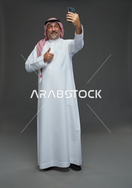 رجل  عربي سعودي خليجي مسن يرتدي الزي الثوب الابيض والشماغ ويمسك الهاتف المحمول, التقاط صورة سيلفي, اجراء مكالمة فيديو عبر الجوال,  ايماءات واشارات مختلفه , كامل الجسم, خلفيه رماديه