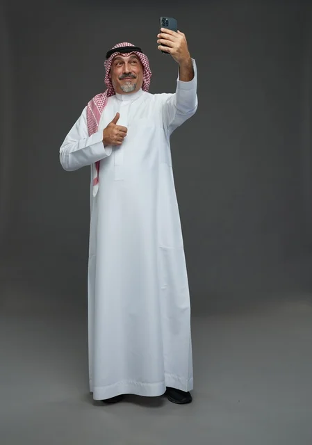 رجل  عربي سعودي خليجي مسن يرتدي الزي الثوب الابيض والشماغ ويمسك الهاتف المحمول, التقاط صورة سيلفي, اجراء مكالمة فيديو عبر الجوال,  ايماءات واشارات مختلفه , كامل الجسم, خلفيه رماديه