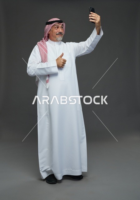 رجل  عربي سعودي خليجي مسن يرتدي الزي الثوب الابيض والشماغ ويمسك الهاتف المحمول, التقاط صورة سيلفي, اجراء مكالمة فيديو عبر الجوال,  ايماءات واشارات مختلفه , كامل الجسم, خلفيه رماديه
