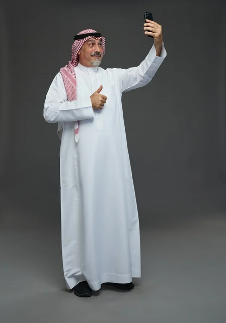 An elderly Arab Saudi Gulf man wearing a white thobe and a shemagh, holding a mobile phone, taking a selfie, making a video call via the phone, with various gestures and signals, full body, gray background.
