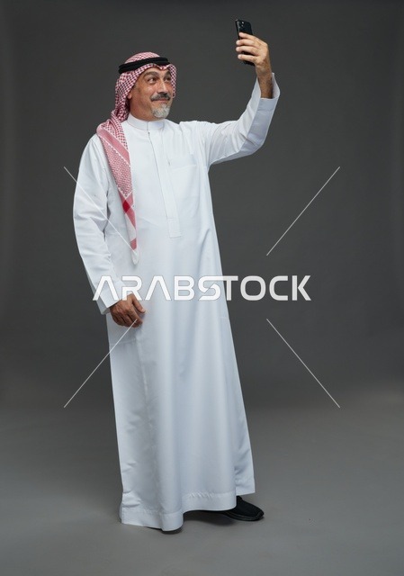 رجل  عربي سعودي خليجي مسن يرتدي الزي الثوب الابيض والشماغ ويمسك الهاتف المحمول, التقاط صورة سيلفي, اجراء مكالمة فيديو عبر الجوال,  ايماءات واشارات مختلفه , كامل الجسم, خلفيه رماديه