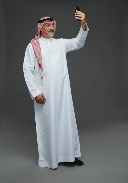 An elderly Arab Saudi Gulf man wearing a white thobe and a shemagh, holding a mobile phone, taking a selfie, making a video call via the phone, with various gestures and signals, full body, gray background.