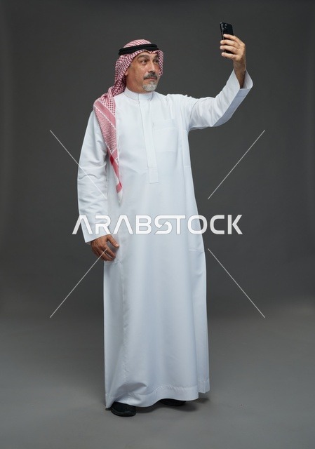 An elderly Arab Saudi Gulf man wearing a white thobe and a shemagh, holding a mobile phone, taking a selfie, making a video call via the phone, with various gestures and signals, full body, gray background.