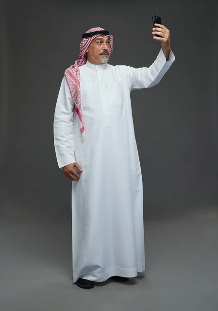 An elderly Arab Saudi Gulf man wearing a white thobe and a shemagh, holding a mobile phone, taking a selfie, making a video call via the phone, with various gestures and signals, full body, gray background.