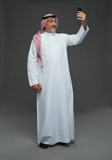 رجل  عربي سعودي خليجي مسن يرتدي الزي الثوب الابيض والشماغ ويمسك الهاتف المحمول, التقاط صورة سيلفي, اجراء مكالمة فيديو عبر الجوال,  ايماءات واشارات مختلفه , كامل الجسم, خلفيه رماديه