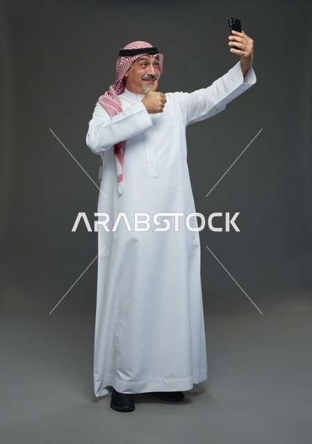 رجل  عربي سعودي خليجي مسن يرتدي الزي الثوب الابيض والشماغ ويمسك الهاتف المحمول, التقاط صورة سيلفي, اجراء مكالمة فيديو عبر الجوال,  ايماءات واشارات مختلفه , كامل الجسم, خلفيه رماديه