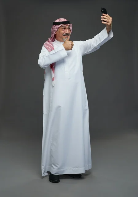 An elderly Arab Saudi Gulf man wearing a white thobe and a shemagh, holding a mobile phone, taking a selfie, making a video call via the phone, with various gestures and signals, full body, gray background.