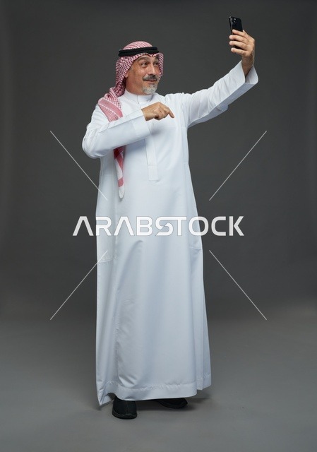 رجل  عربي سعودي خليجي مسن يرتدي الزي الثوب الابيض والشماغ ويمسك الهاتف المحمول, التقاط صورة سيلفي, اجراء مكالمة فيديو عبر الجوال,  ايماءات واشارات مختلفه , كامل الجسم, خلفيه رماديه