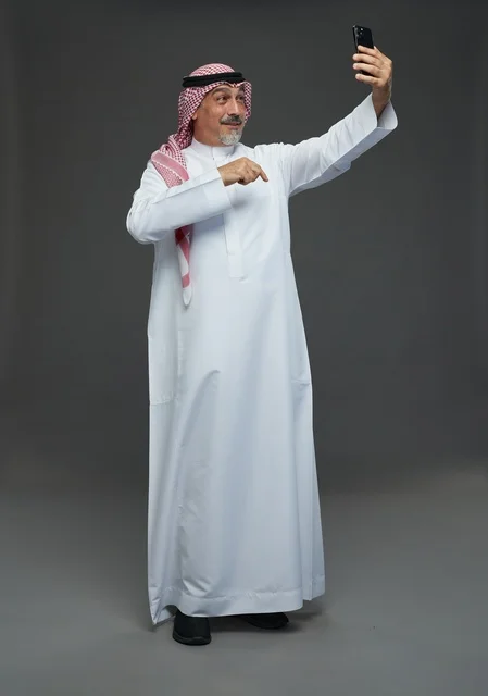 An elderly Arab Saudi Gulf man wearing a white thobe and a shemagh, holding a mobile phone, taking a selfie, making a video call via the phone, with various gestures and signals, full body, gray background.