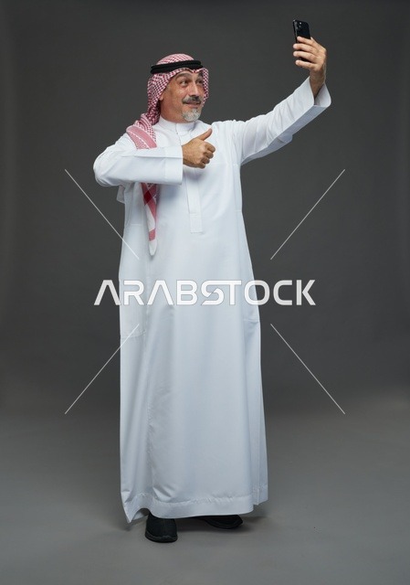 An elderly Arab Saudi Gulf man wearing a white thobe and a shemagh, holding a mobile phone, taking a selfie, making a video call via the phone, with various gestures and signals, full body, gray background.