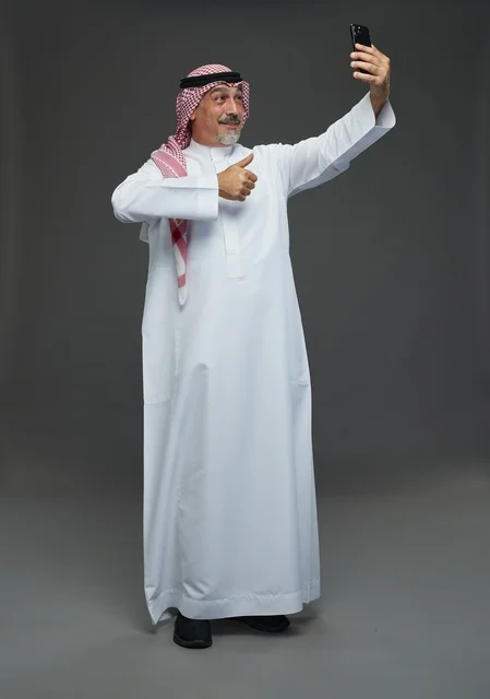 An elderly Arab Saudi Gulf man wearing a white thobe and a shemagh, holding a mobile phone, taking a selfie, making a video call via the phone, with various gestures and signals, full body, gray background.
