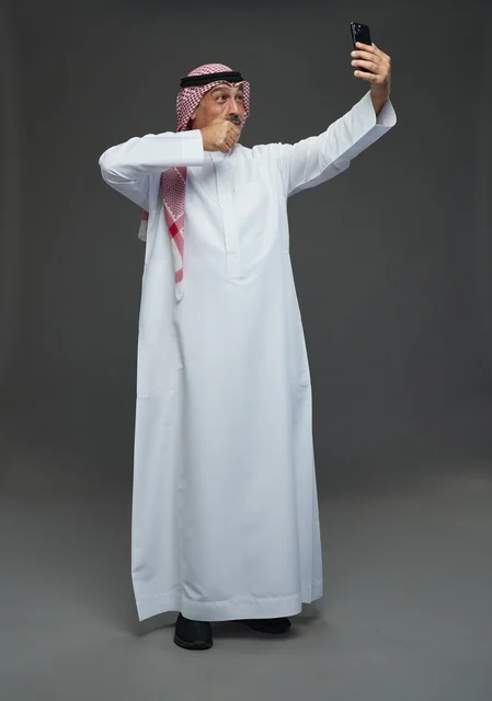 رجل  عربي سعودي خليجي مسن يرتدي الزي الثوب الابيض والشماغ ويمسك الهاتف المحمول, التقاط صورة سيلفي, اجراء مكالمة فيديو عبر الجوال,  ايماءات واشارات مختلفه , كامل الجسم, خلفيه رماديه