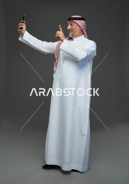 رجل  عربي سعودي خليجي مسن يرتدي الزي الثوب الابيض والشماغ ويمسك الهاتف المحمول, التقاط صورة سيلفي, اجراء مكالمة فيديو عبر الجوال,  ايماءات واشارات مختلفه , كامل الجسم, خلفيه رماديه