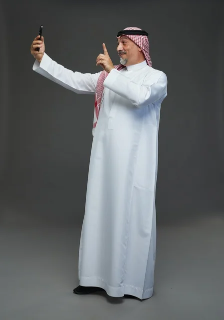 An elderly Arab Saudi Gulf man wearing a white thobe and a shemagh, holding a mobile phone, taking a selfie, making a video call via the phone, with various gestures and signals, full body, gray background.