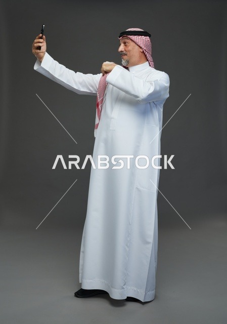 رجل  عربي سعودي خليجي مسن يرتدي الزي الثوب الابيض والشماغ ويمسك الهاتف المحمول, التقاط صورة سيلفي, اجراء مكالمة فيديو عبر الجوال,  ايماءات واشارات مختلفه , كامل الجسم, خلفيه رماديه
