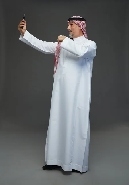 رجل  عربي سعودي خليجي مسن يرتدي الزي الثوب الابيض والشماغ ويمسك الهاتف المحمول, التقاط صورة سيلفي, اجراء مكالمة فيديو عبر الجوال,  ايماءات واشارات مختلفه , كامل الجسم, خلفيه رماديه