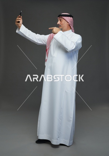 رجل  عربي سعودي خليجي مسن يرتدي الزي الثوب الابيض والشماغ ويمسك الهاتف المحمول, التقاط صورة سيلفي, اجراء مكالمة فيديو عبر الجوال,  ايماءات واشارات مختلفه , كامل الجسم, خلفيه رماديه