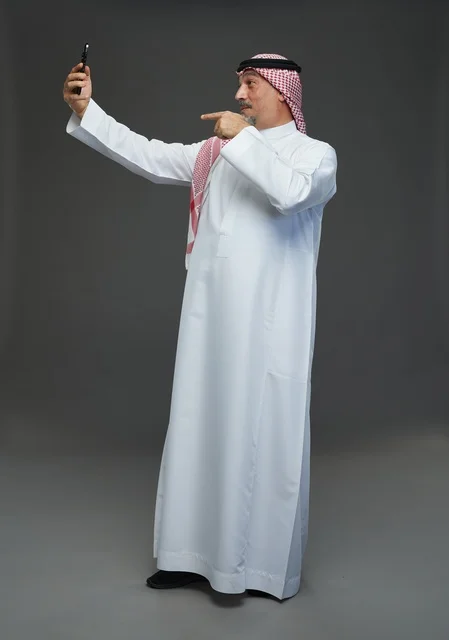 An elderly Arab Saudi Gulf man wearing a white thobe and a shemagh, holding a mobile phone, taking a selfie, making a video call via the phone, with various gestures and signals, full body, gray background.