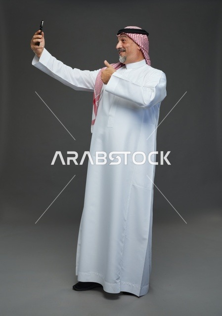 رجل  عربي سعودي خليجي مسن يرتدي الزي الثوب الابيض والشماغ ويمسك الهاتف المحمول, التقاط صورة سيلفي, اجراء مكالمة فيديو عبر الجوال,  ايماءات واشارات مختلفه , كامل الجسم, خلفيه رماديه