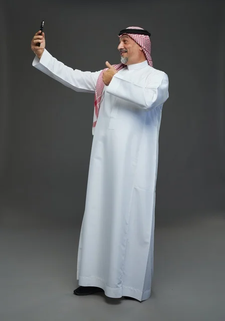 رجل  عربي سعودي خليجي مسن يرتدي الزي الثوب الابيض والشماغ ويمسك الهاتف المحمول, التقاط صورة سيلفي, اجراء مكالمة فيديو عبر الجوال,  ايماءات واشارات مختلفه , كامل الجسم, خلفيه رماديه