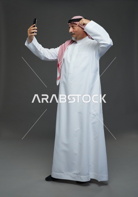 رجل  عربي سعودي خليجي مسن يرتدي الزي الثوب الابيض والشماغ ويمسك الهاتف المحمول, التقاط صورة سيلفي, اجراء مكالمة فيديو عبر الجوال,  ايماءات واشارات مختلفه , كامل الجسم, خلفيه رماديه