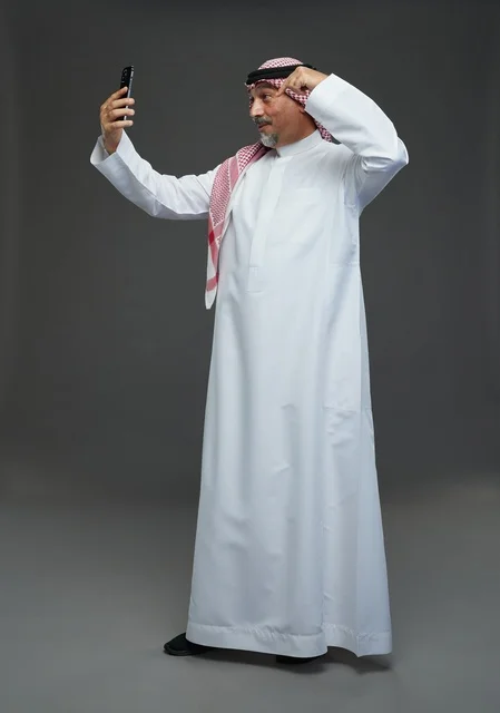An elderly Arab Saudi Gulf man wearing a white thobe and a shemagh, holding a mobile phone, taking a selfie, making a video call via the phone, with various gestures and signals, full body, gray background.