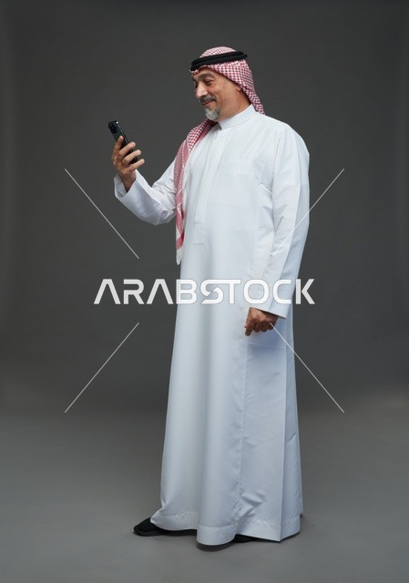 رجل  عربي سعودي خليجي مسن يرتدي الزي الثوب الابيض والشماغ ويمسك الهاتف المحمول, التقاط صورة سيلفي, اجراء مكالمة فيديو عبر الجوال,  ايماءات واشارات مختلفه , كامل الجسم, خلفيه رماديه