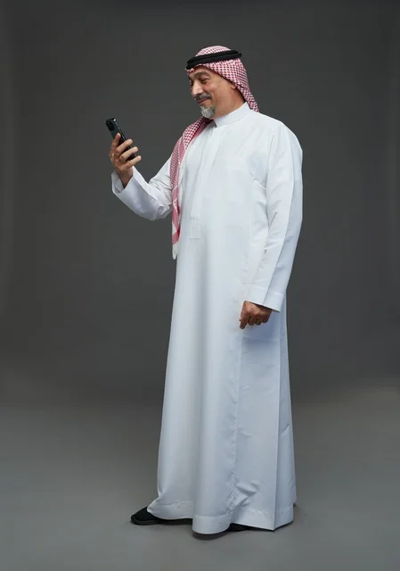 رجل  عربي سعودي خليجي مسن يرتدي الزي الثوب الابيض والشماغ ويمسك الهاتف المحمول, التقاط صورة سيلفي, اجراء مكالمة فيديو عبر الجوال,  ايماءات واشارات مختلفه , كامل الجسم, خلفيه رماديه