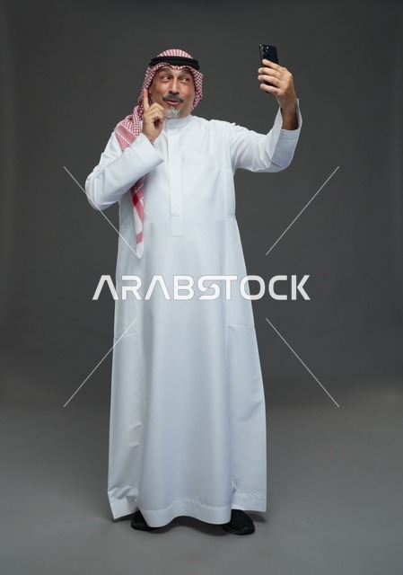 رجل  عربي سعودي خليجي مسن يرتدي الزي الثوب الابيض والشماغ ويمسك الهاتف المحمول, التقاط صورة سيلفي, اجراء مكالمة فيديو عبر الجوال,  ايماءات واشارات مختلفه , كامل الجسم, خلفيه رماديه
