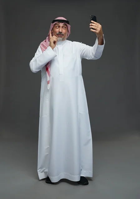 An elderly Arab Saudi Gulf man wearing a white thobe and a shemagh, holding a mobile phone, taking a selfie, making a video call via the phone, with various gestures and signals, full body, gray background.