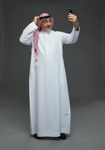 An elderly Arab Saudi Gulf man wearing a white thobe and a shemagh, holding a mobile phone, taking a selfie, making a video call via the phone, with various gestures and signals, full body, gray background.