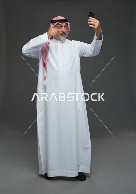 رجل  عربي سعودي خليجي مسن يرتدي الزي الثوب الابيض والشماغ ويمسك الهاتف المحمول, التقاط صورة سيلفي, اجراء مكالمة فيديو عبر الجوال,  ايماءات واشارات مختلفه , كامل الجسم, خلفيه رماديه