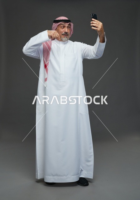 رجل  عربي سعودي خليجي مسن يرتدي الزي الثوب الابيض والشماغ ويمسك الهاتف المحمول, التقاط صورة سيلفي, اجراء مكالمة فيديو عبر الجوال,  ايماءات واشارات مختلفه , كامل الجسم, خلفيه رماديه