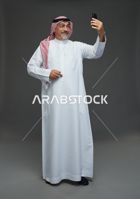 An elderly Arab Saudi Gulf man wearing a white thobe and a shemagh, holding a mobile phone, taking a selfie, making a video call via the phone, with various gestures and signals, full body, gray background.