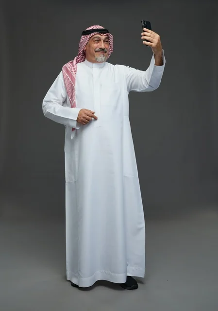 رجل  عربي سعودي خليجي مسن يرتدي الزي الثوب الابيض والشماغ ويمسك الهاتف المحمول, التقاط صورة سيلفي, اجراء مكالمة فيديو عبر الجوال,  ايماءات واشارات مختلفه , كامل الجسم, خلفيه رماديه